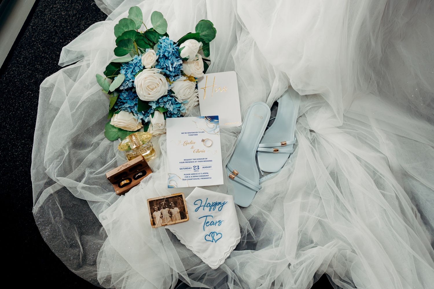 Wedding details including blue and white flowers, invitation, and veil arranged artistically on dark surface.