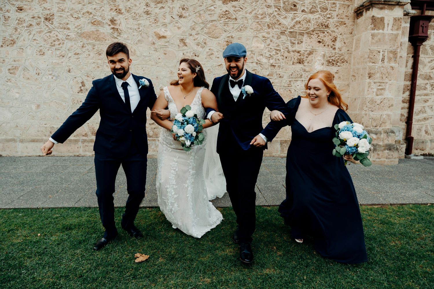 Wedding party in formal attire walks arm in arm and laughs together on grass near stone wall.