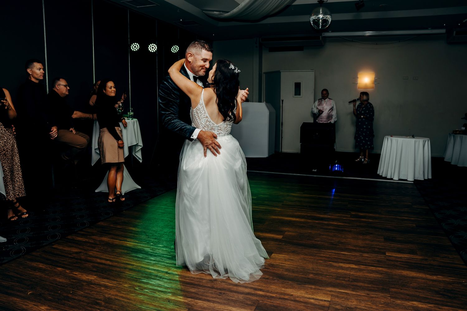 A couple performs their first dance at their wedding reception on a wooden dance floor under dim lighting with green accents.