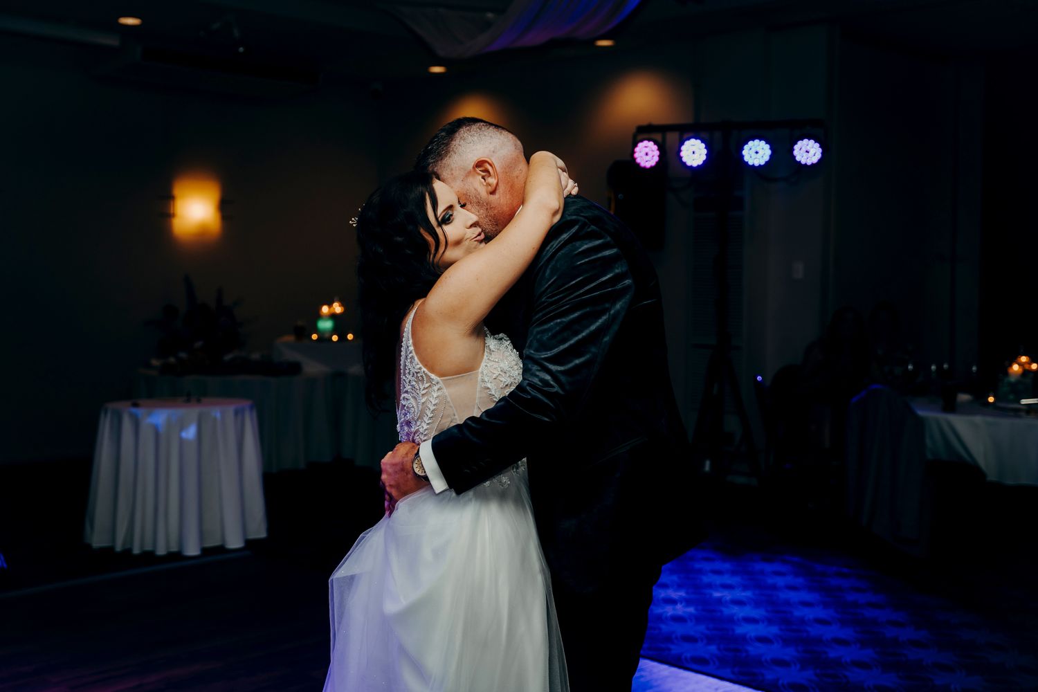 A couple shares a romantic dance moment at their wedding reception with blue mood lighting in the background.