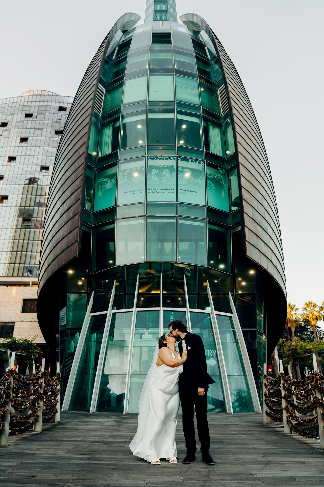 A couple embraces in front of a modern glass building with teal-tinted windows and curved architectural design.
