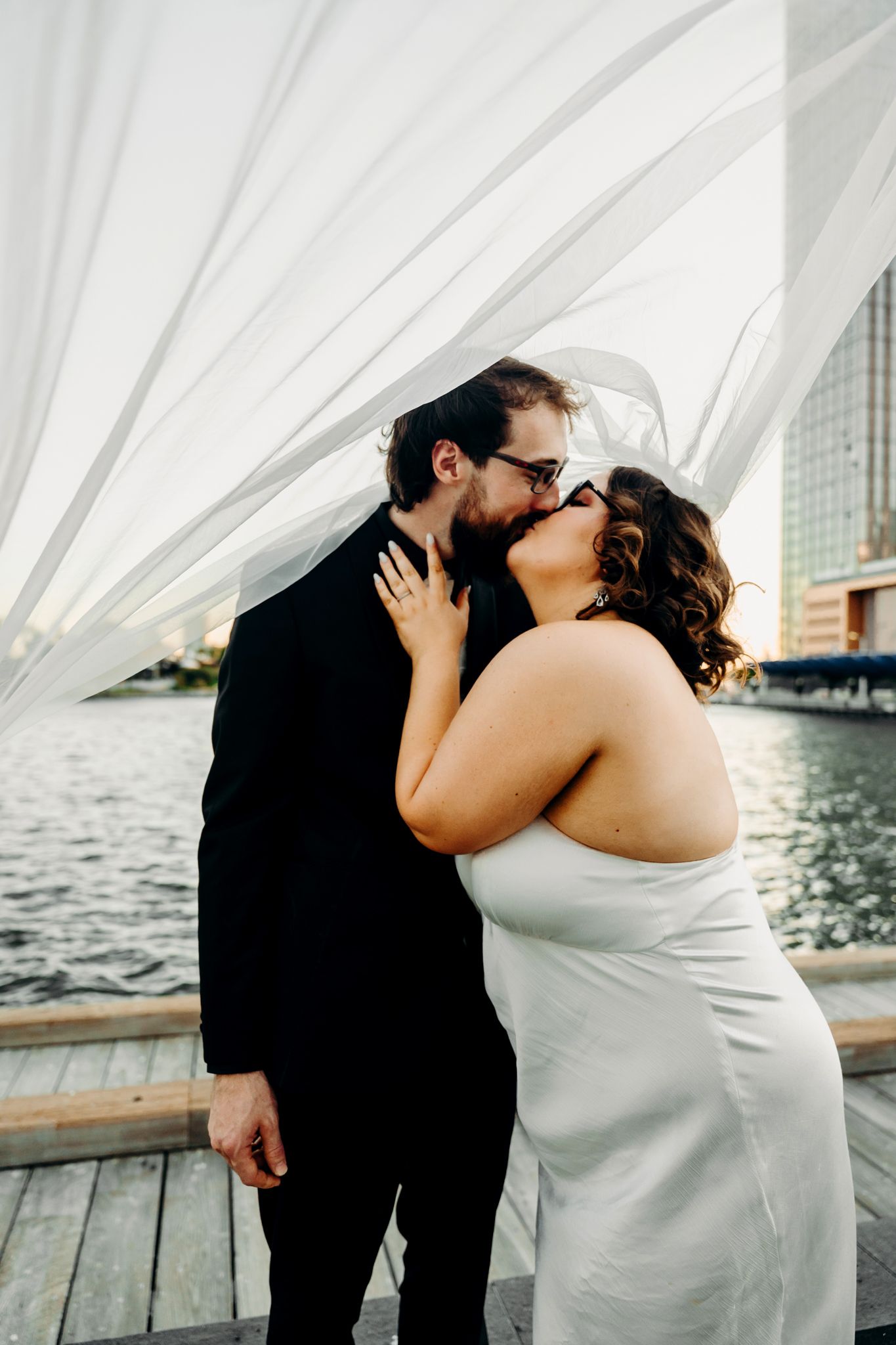 A romantic wedding couple sharing a kiss on a waterfront deck with flowing white fabric and water in background.