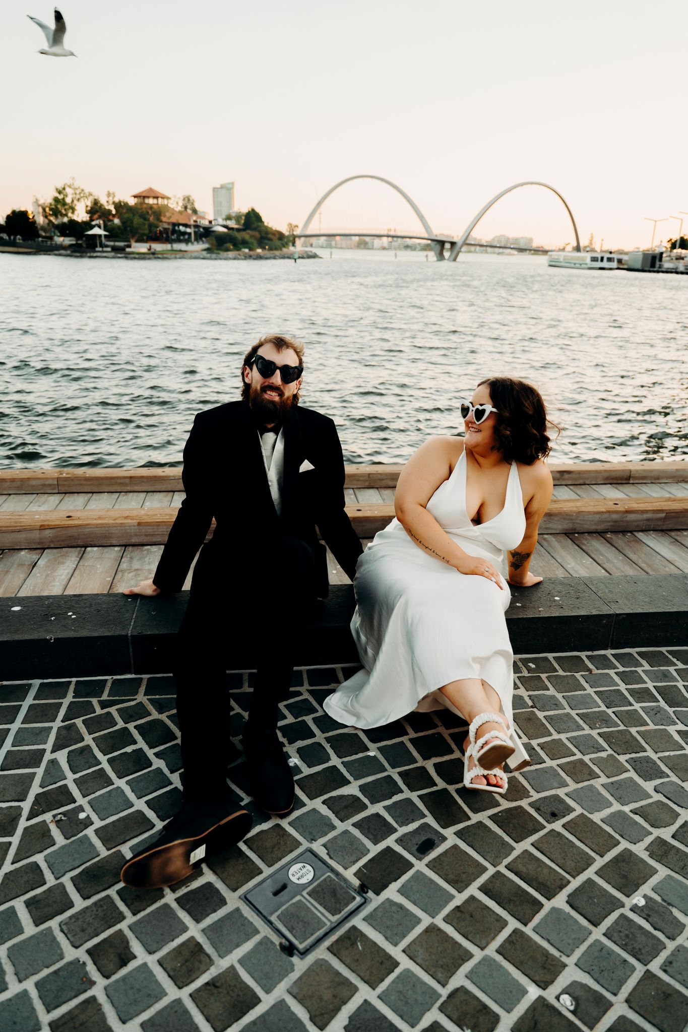 A couple sits on a waterfront walkway at sunset with a distinctive arched bridge visible across the water.