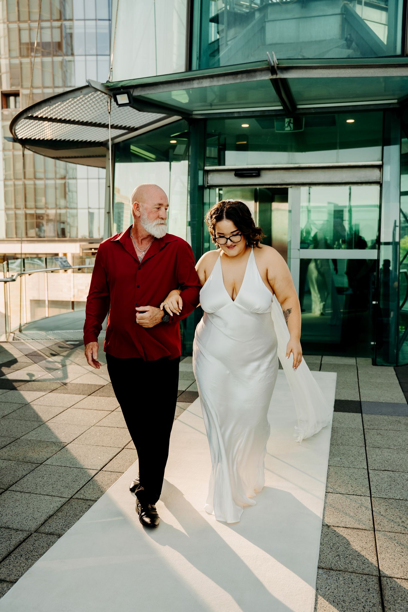 Two people in formal attire walk together on a tiled outdoor terrace with glass walls in the background.