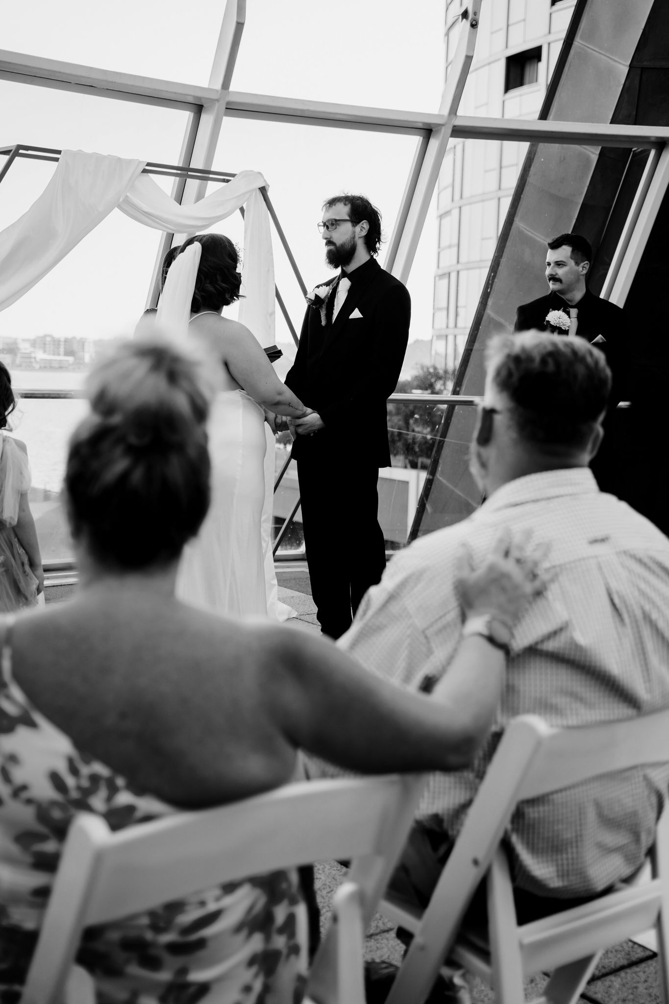 Black and white wedding ceremony photo showing guests seated at an indoor venue with natural light streaming through windows.