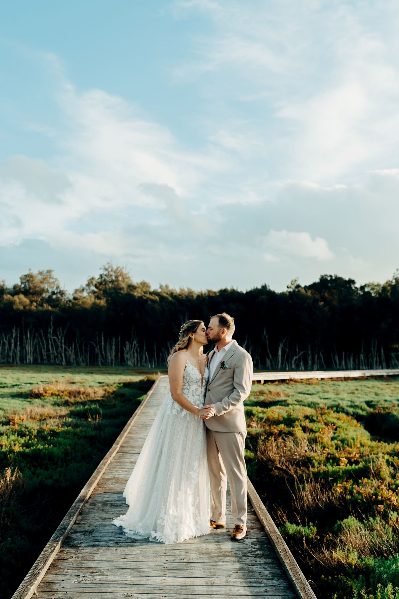 A couple shares an intimate moment on a wooden boardwalk surrounded by marsh grass under a bright blue sky.