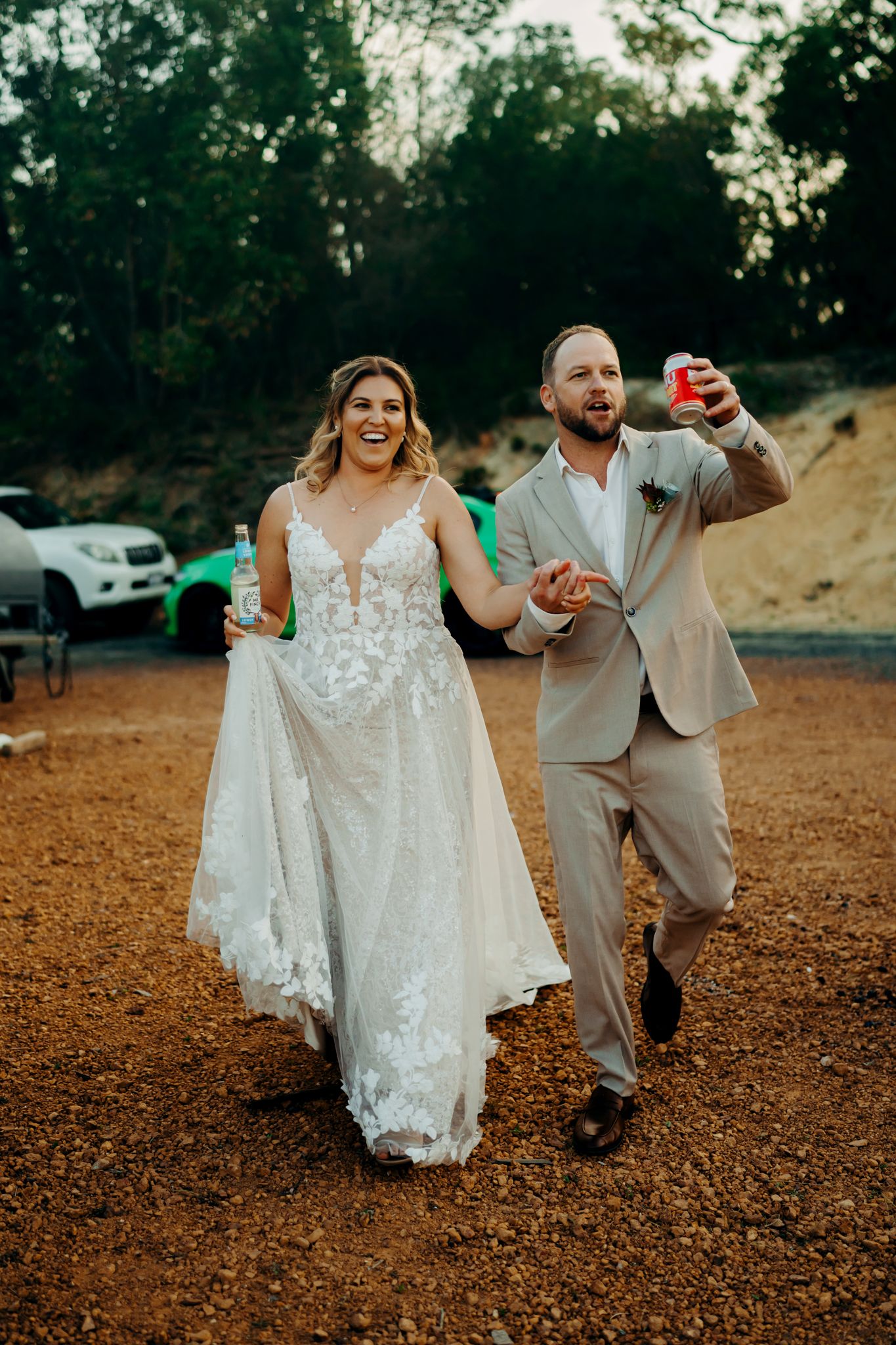 A couple in wedding attire walks together on rustic ground during sunset in a natural outdoor setting.