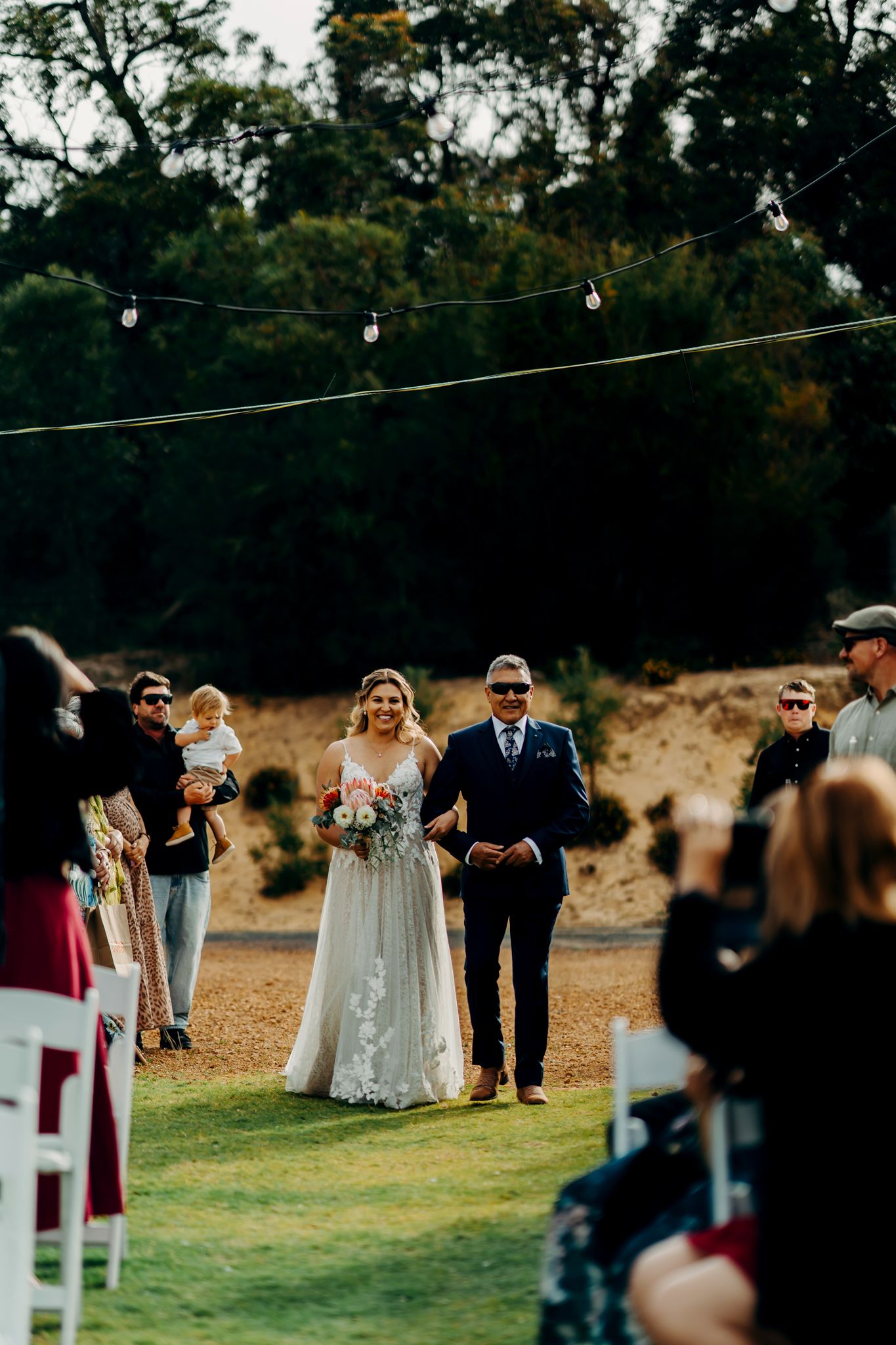 Wedding guests gather on a grassy lawn during an outdoor ceremony with string lights overhead.