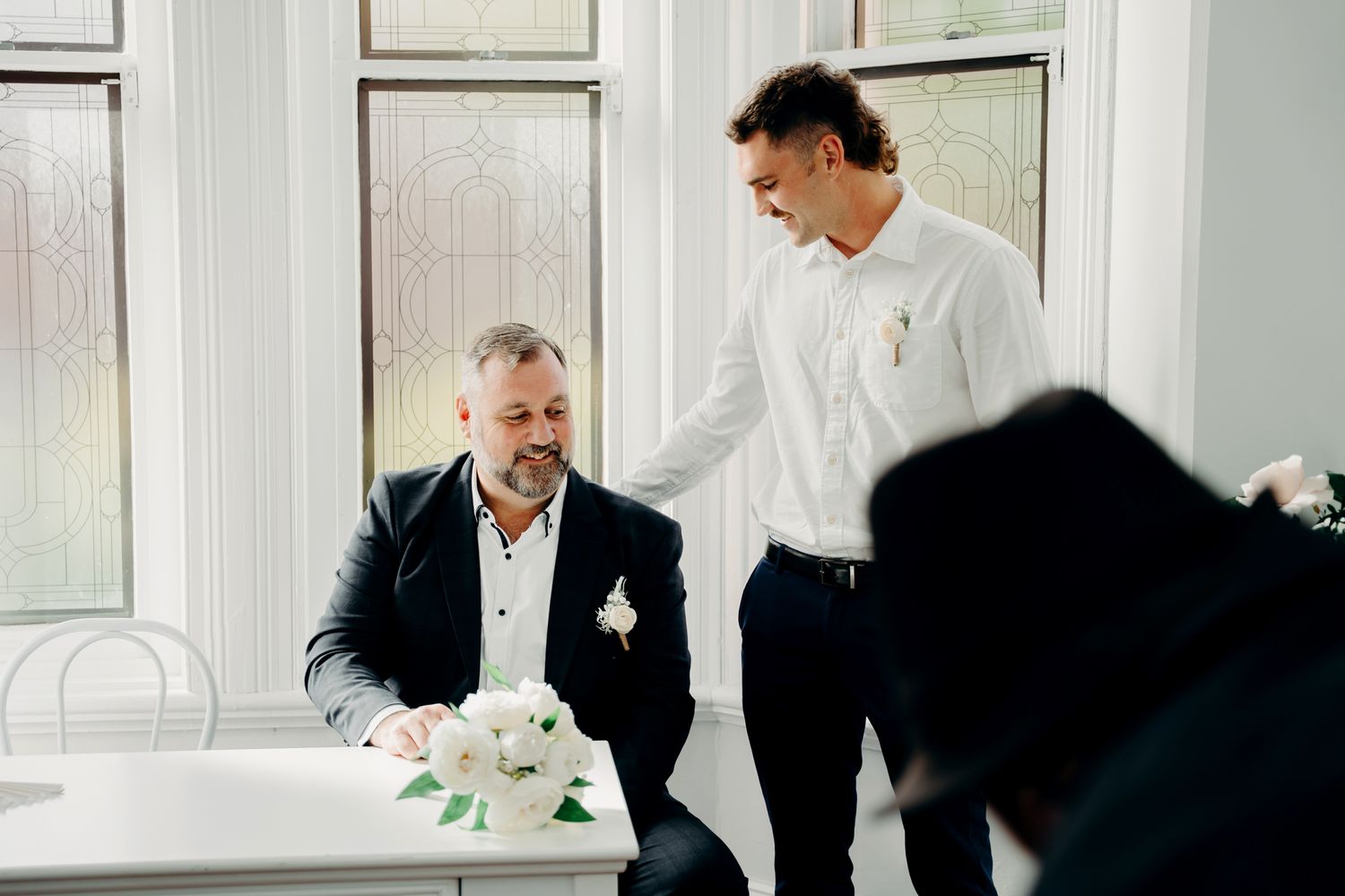 Wedding guests in elegant attire have a conversation at a table decorated with white floral centerpieces.