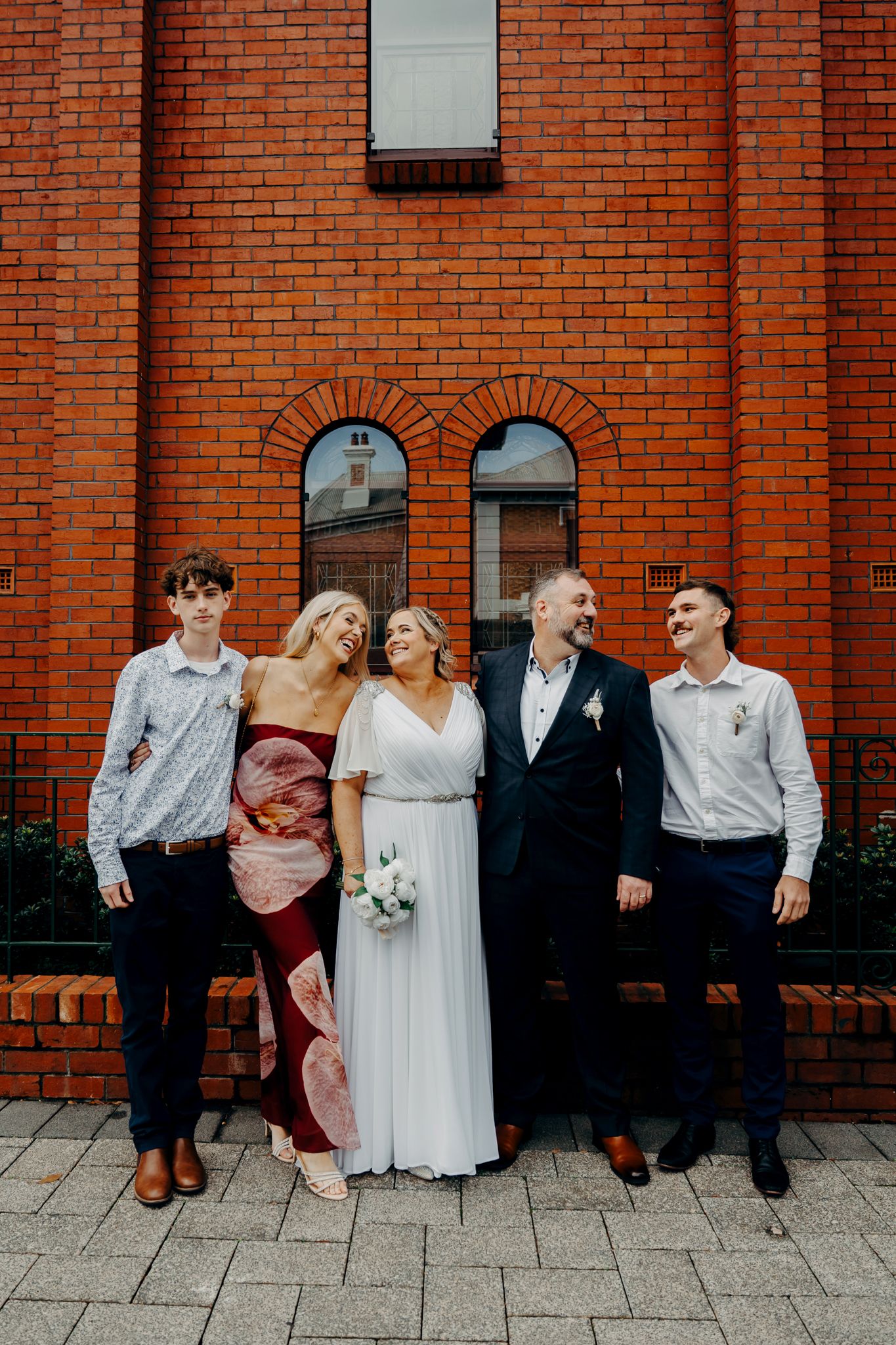 A group stands together for a wedding photo against a rustic red brick building with arched windows.