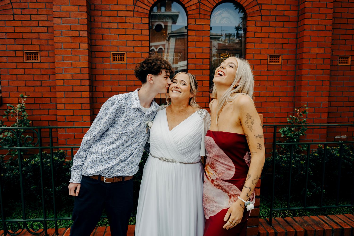 Three people laugh together in front of a red brick building with arched windows.