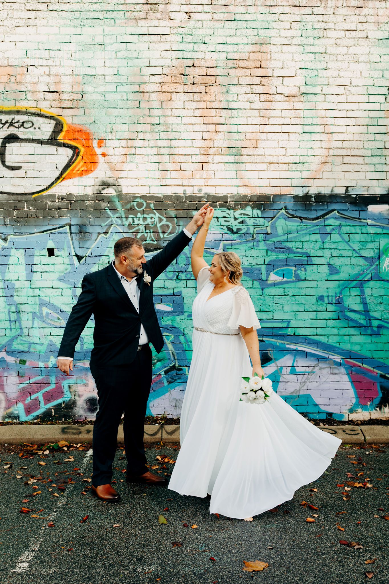 A couple in wedding attire share a joyful moment dancing in front of a colorful graffiti wall.
