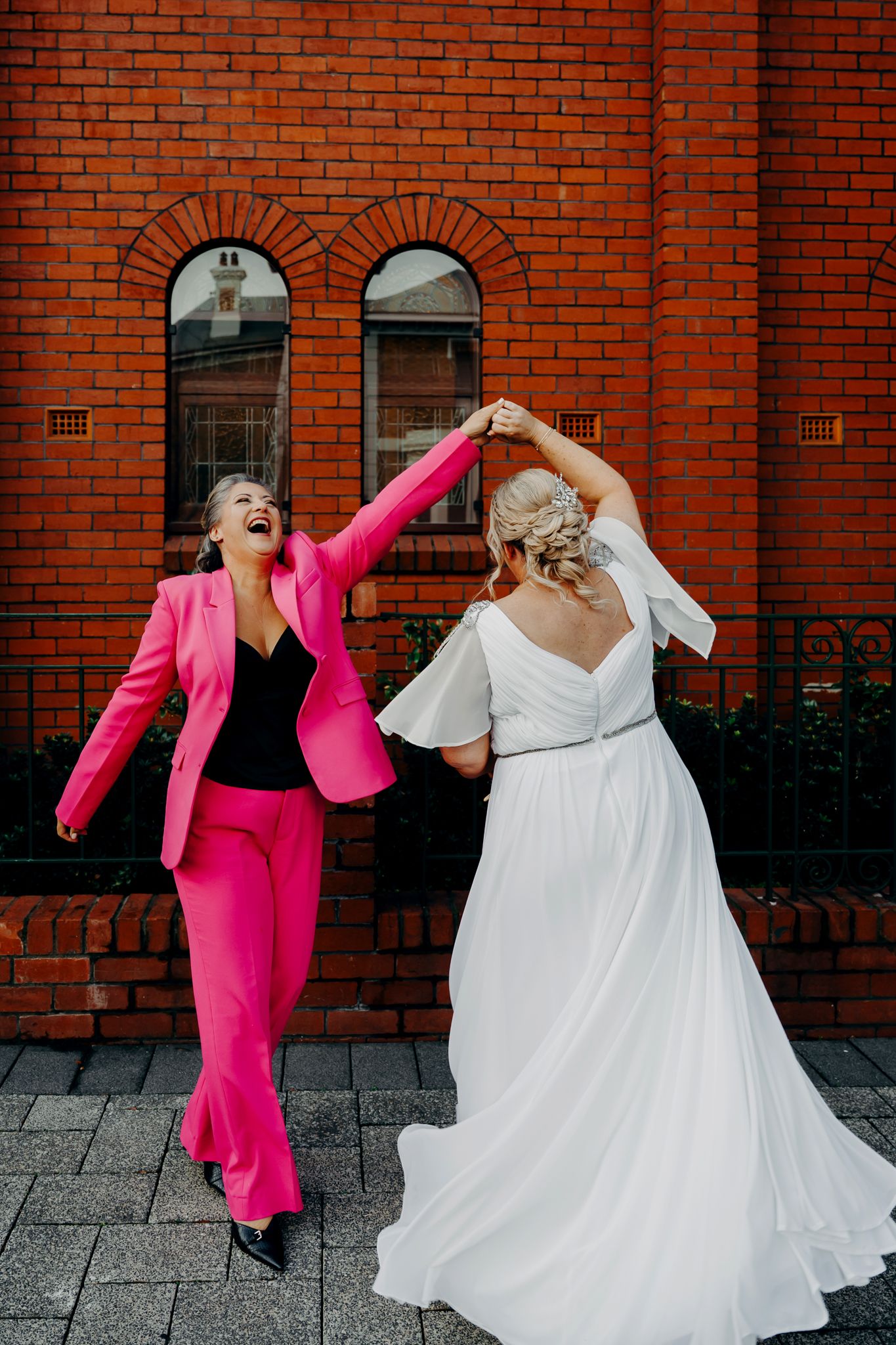Two people dancing joyfully outside a red brick building, one in a hot pink suit and one in a flowing white dress.