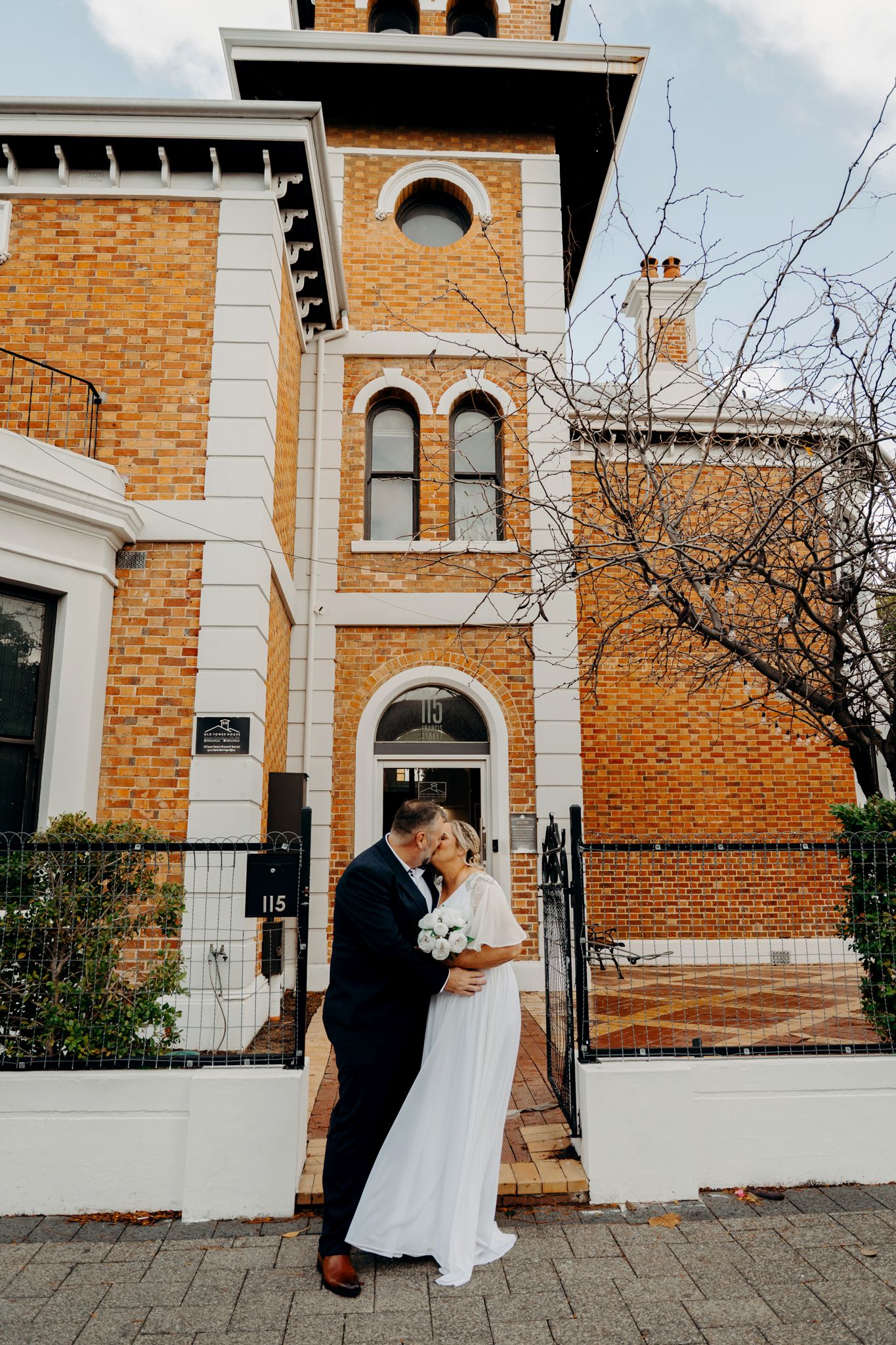 Elegant wedding couple poses in front of historic brick building with white trim architectural details.