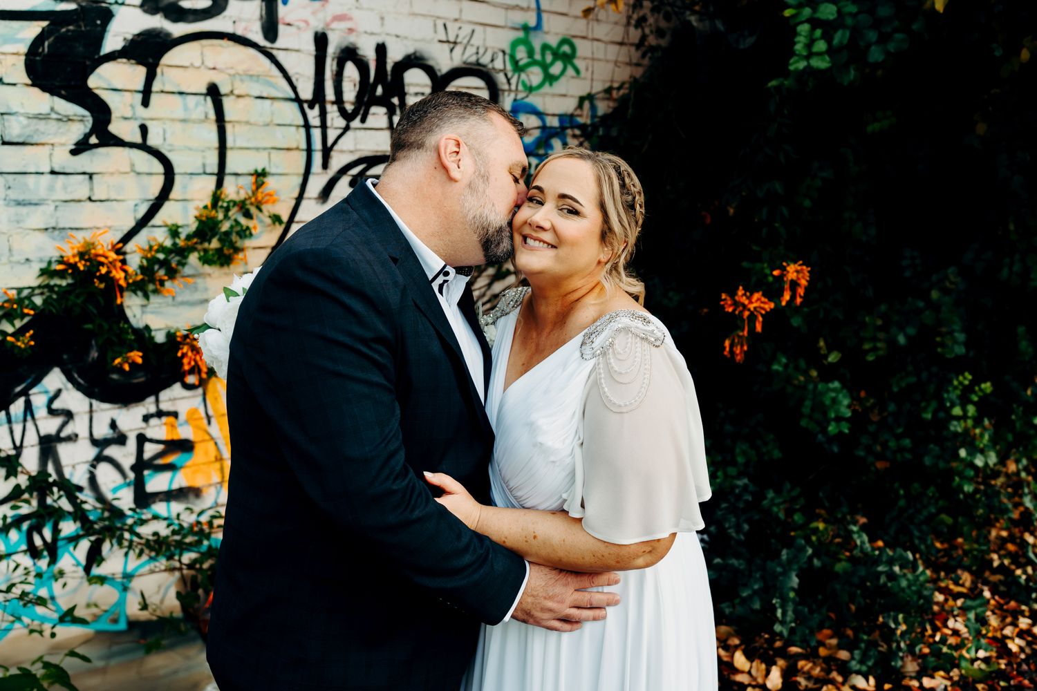 A romantic wedding couple embraces against a graffiti wall with colorful flowers in the background.