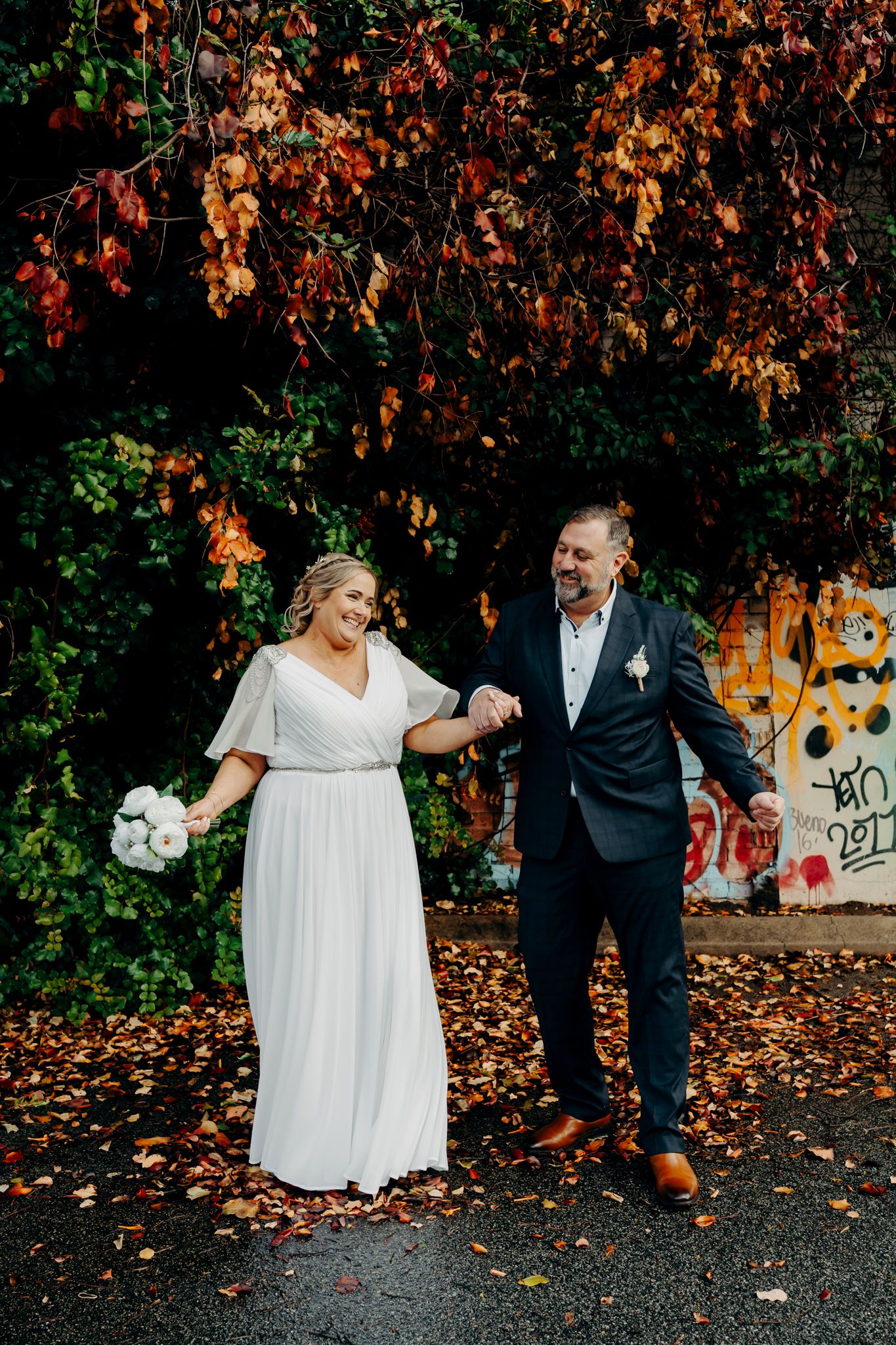 A couple in wedding attire holds hands while standing under autumn trees with orange leaves and a graffiti wall backdrop.