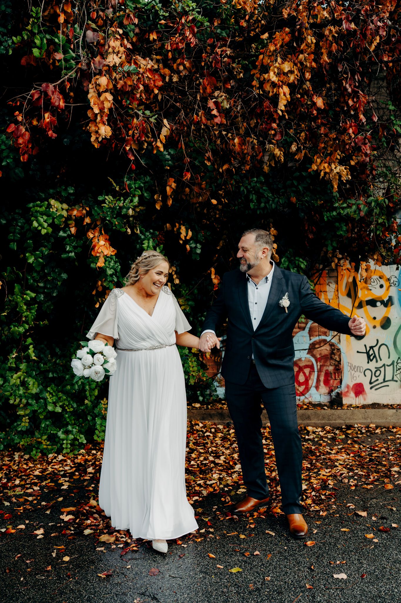 Newlyweds dancing together in autumn setting with fallen leaves and graffiti wall backdrop.
