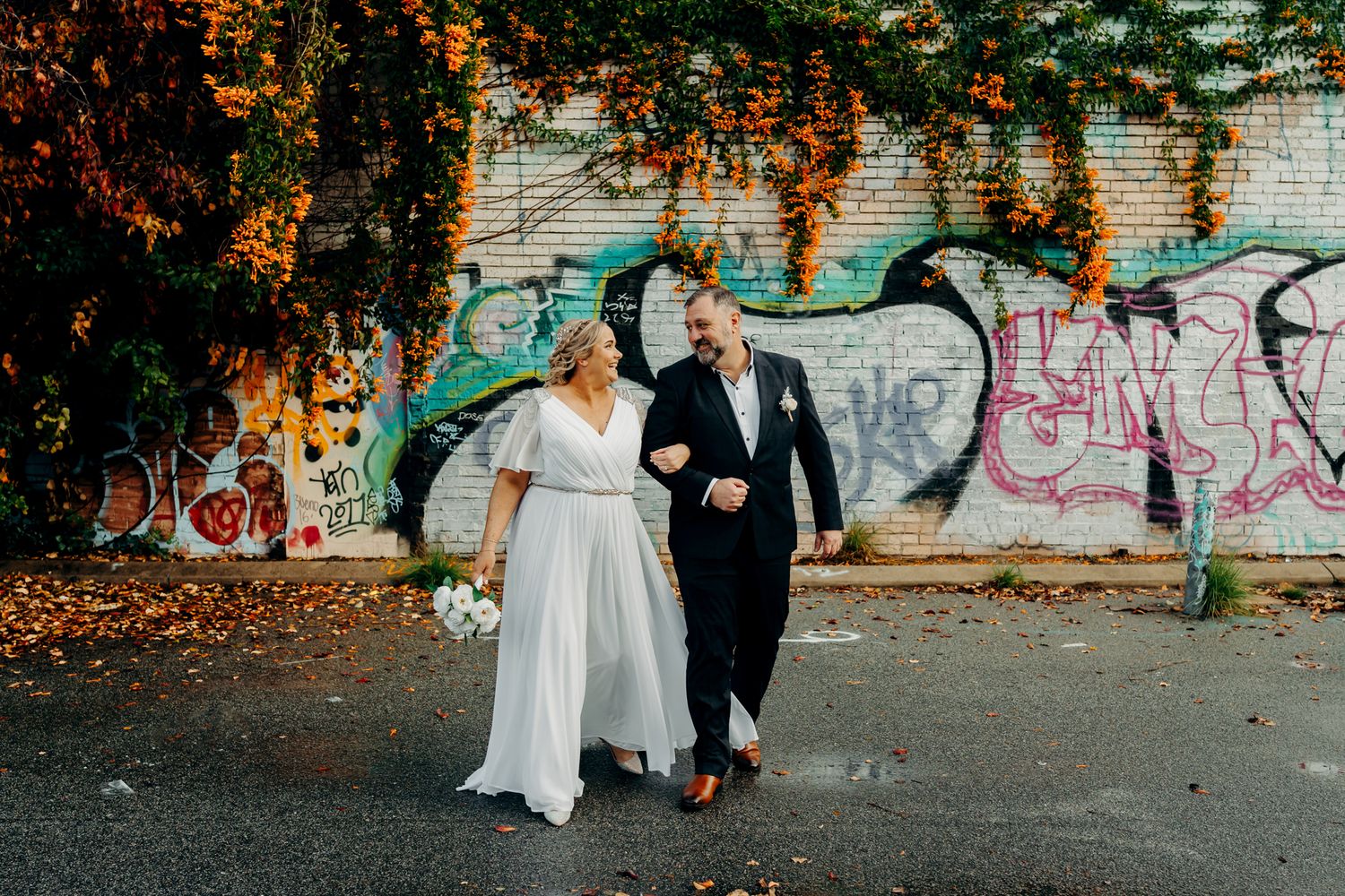 A couple walks together in front of a graffiti-covered brick wall with autumn foliage overhead.