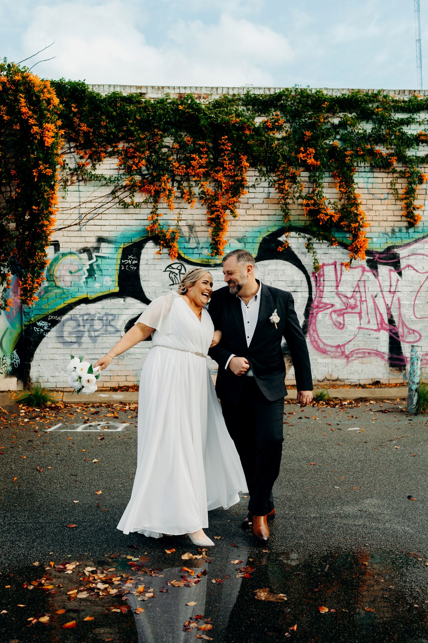 Newly married couple walking together in front of graffiti wall with orange flowers cascading down brick wall.