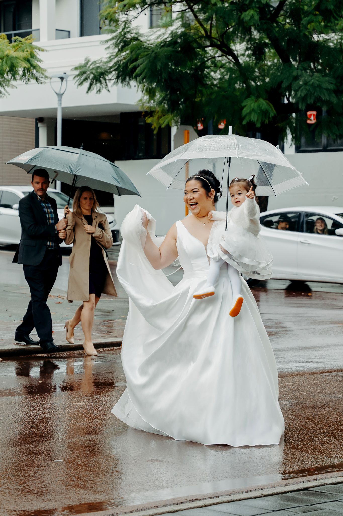 Wedding party walks down rainy city street with umbrellas while bride's white dress flows in the wind.