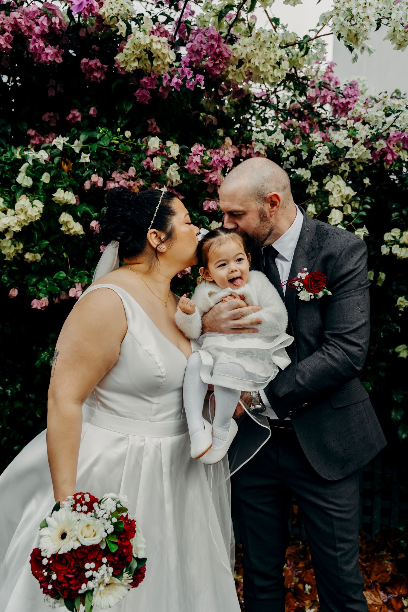Heartwarming family portrait against flowering bougainvillea backdrop with wedding bouquet of red and white flowers.