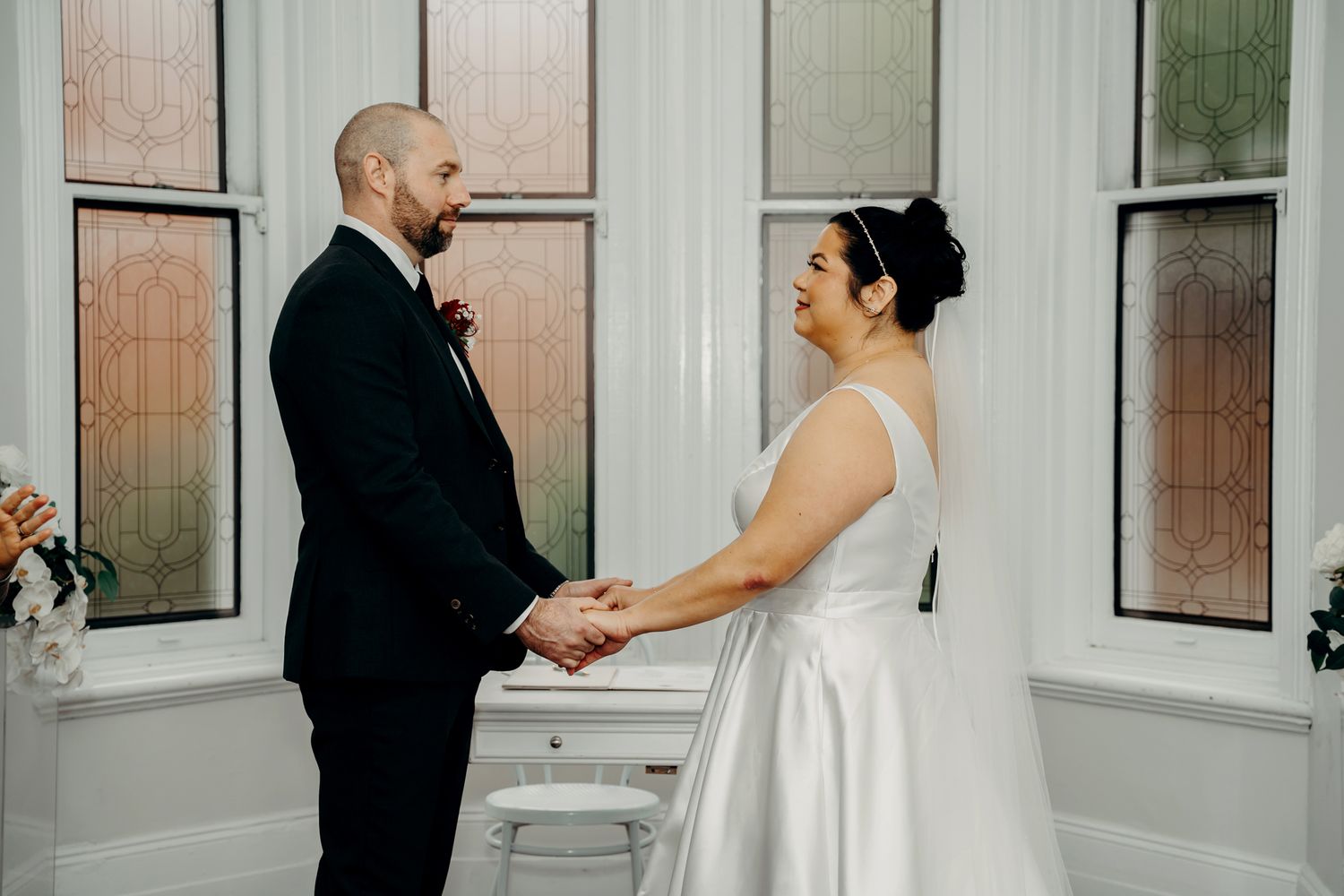 A couple holds hands during an intimate ceremony in a bright window-lit room, wearing formal black and white wedding attire.
