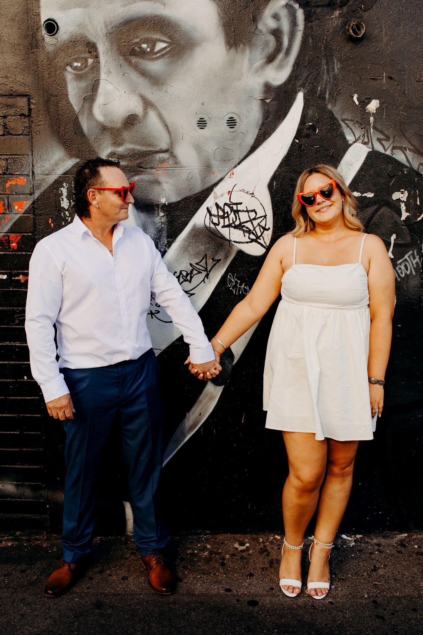 A couple in white outfits holds hands while standing against an urban wall with black and white street art.