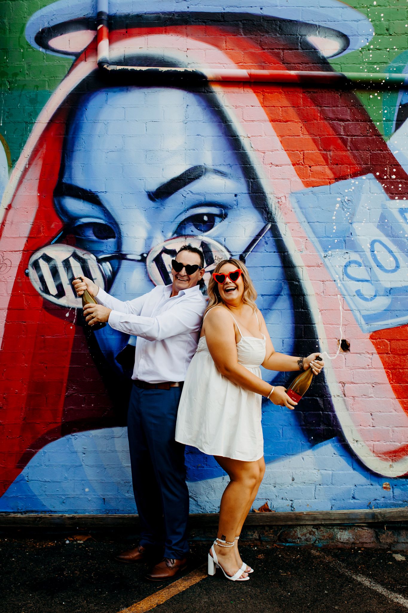 Woman in white dress poses playfully against vibrant street art mural backdrop with colorful geometric designs.