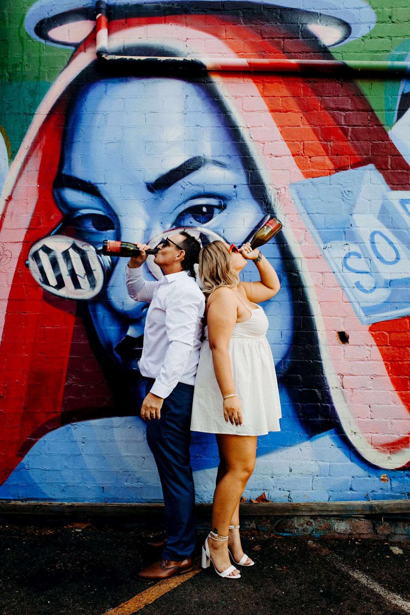 A couple poses playfully against a colorful street art mural on a brick wall in an urban setting.