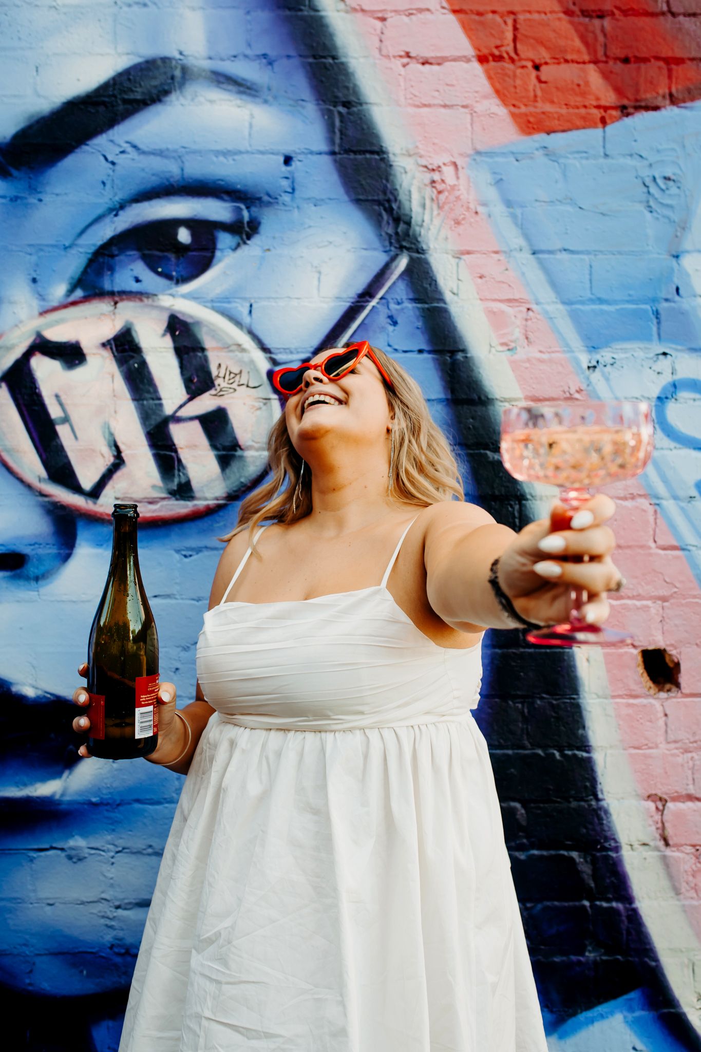 A person in a white sundress laughs joyfully while holding drinks against a colorful graffiti mural background.