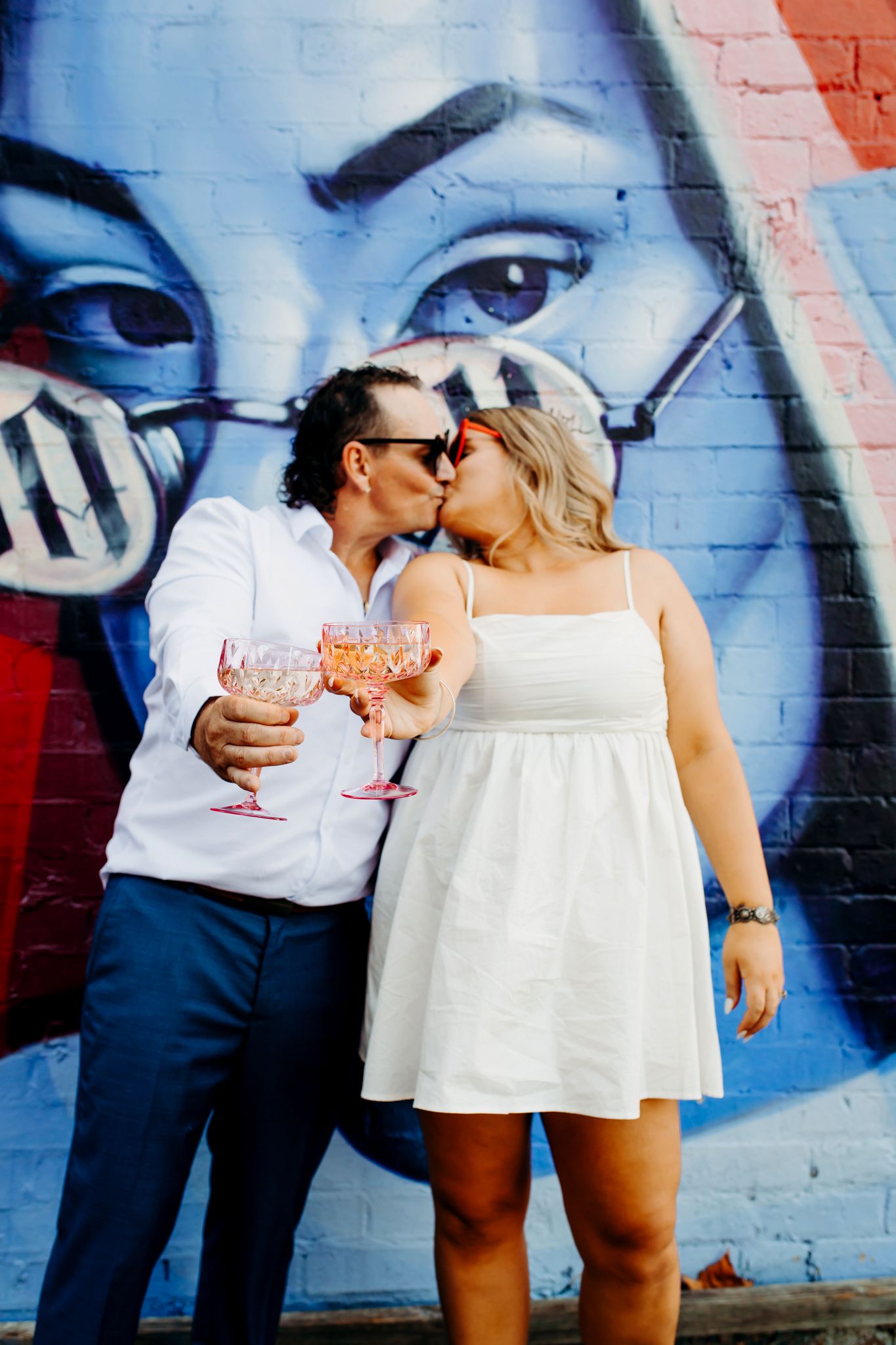 Couple sharing drinks and a kiss against vibrant blue street art mural on brick wall.