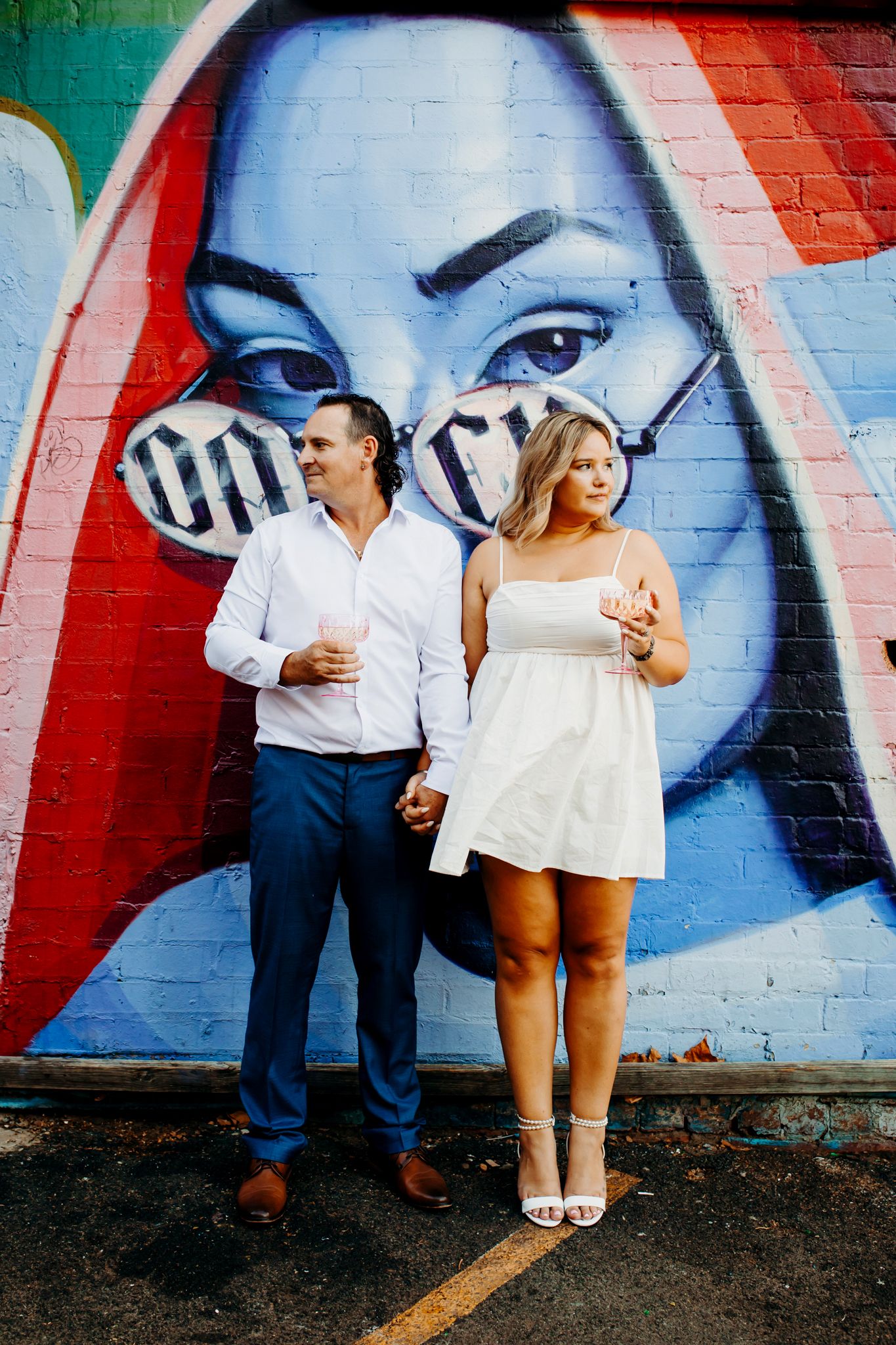 A couple poses against a vibrant street art mural, dressed in white clothing for a stylish engagement photo.