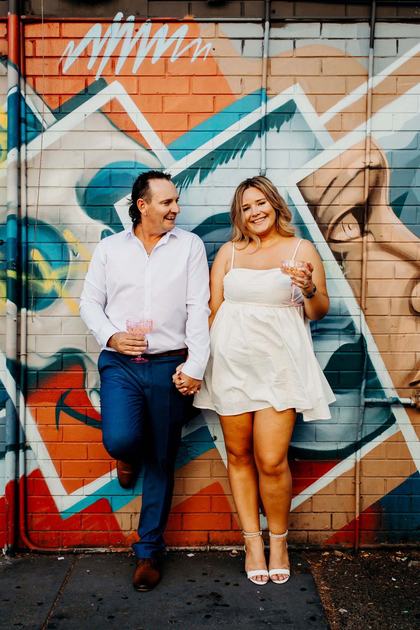 A couple in casual attire laughs together while leaning against a colorful graffiti wall in an urban setting.