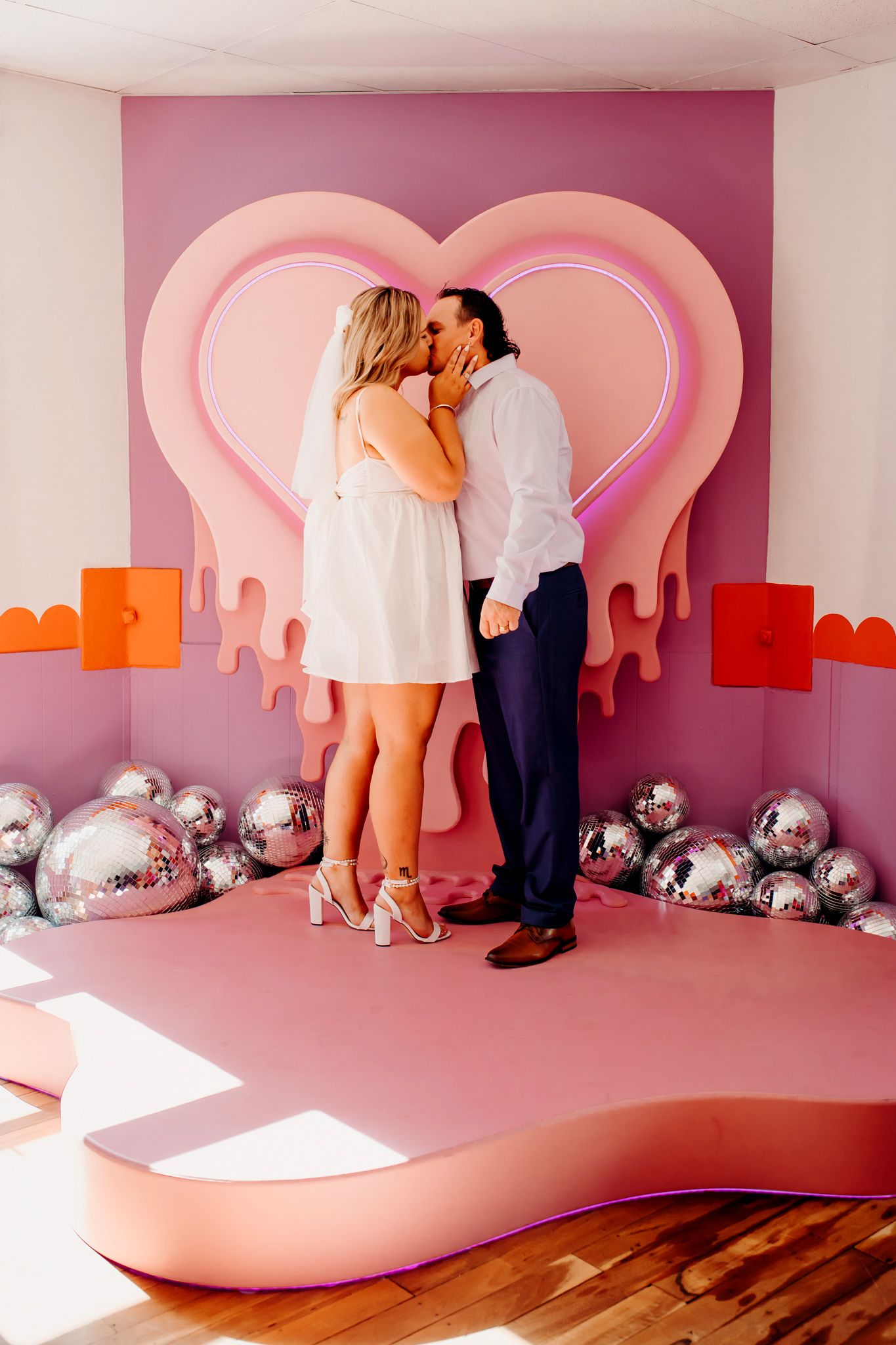 Couple sharing romantic moment in pink room with heart backdrop and silver disco balls.