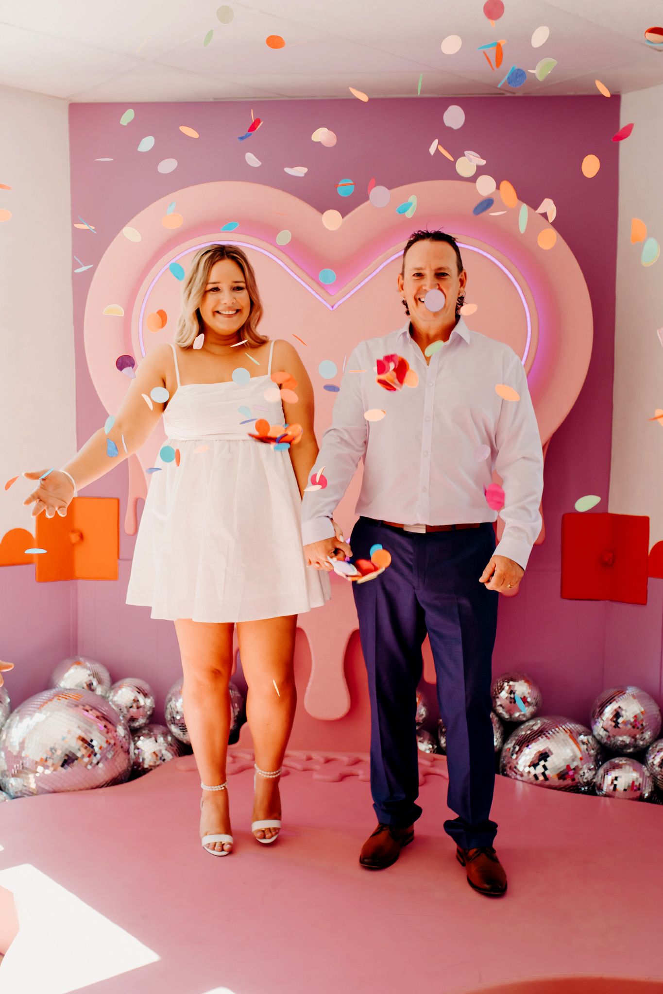 A couple poses in front of a pink heart backdrop with confetti and silver balloons for a festive Valentine's Day photoshoot.