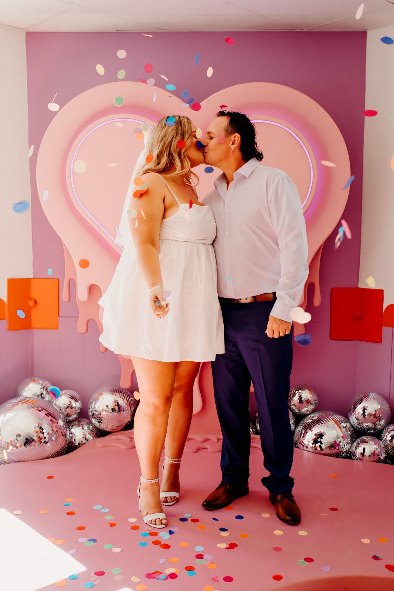 A romantic Valentine's day photoshoot setup with pink heart backdrop, silver disco balls and colorful confetti on the floor.