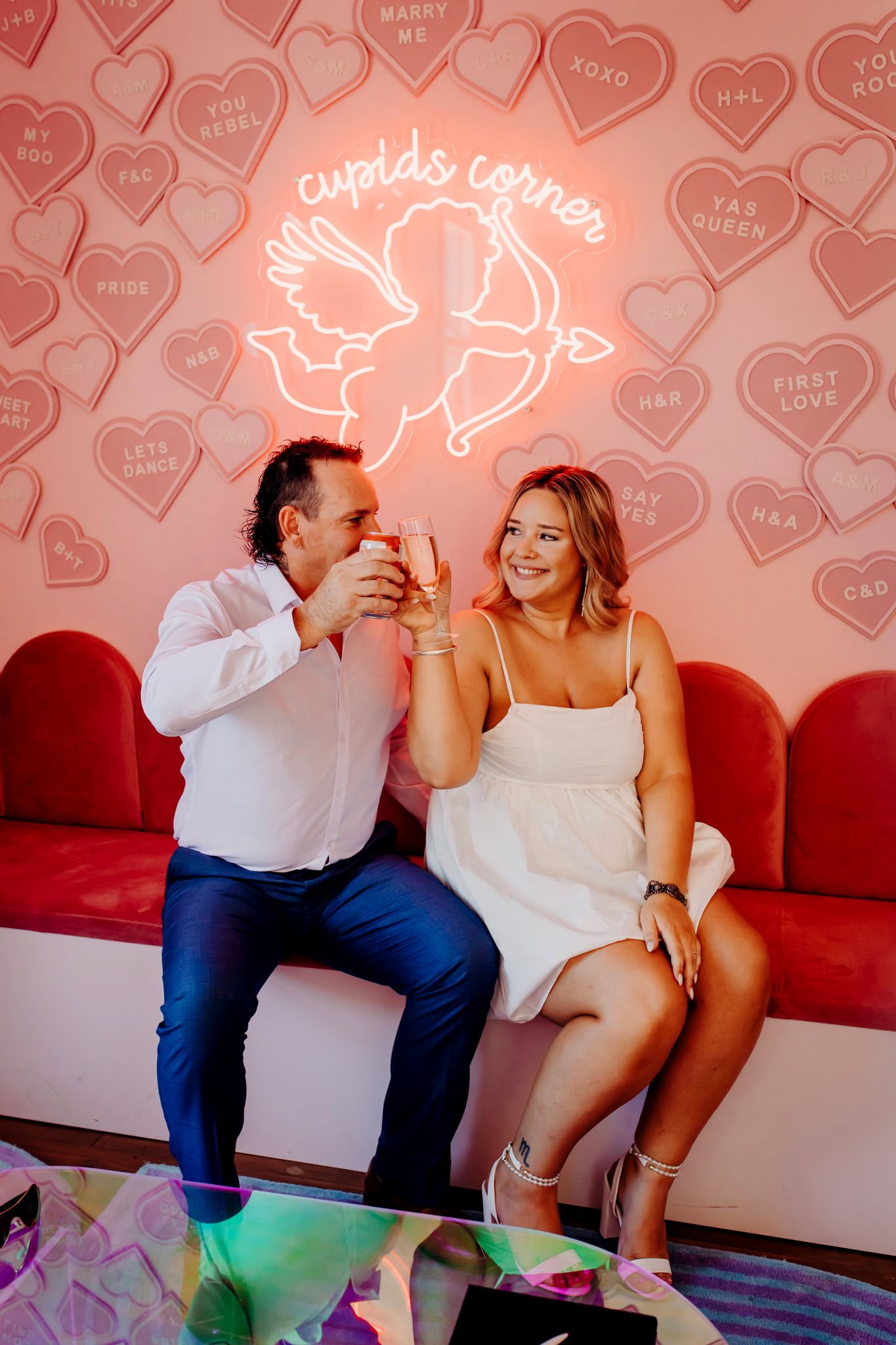 Couple enjoying drinks under neon Cupid sign with pink heart pattern background.