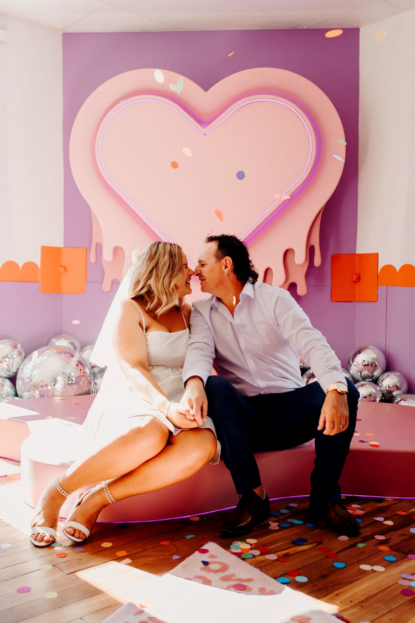 Romantic couple sitting in front of large pink heart backdrop with disco balls and confetti on wooden floor.