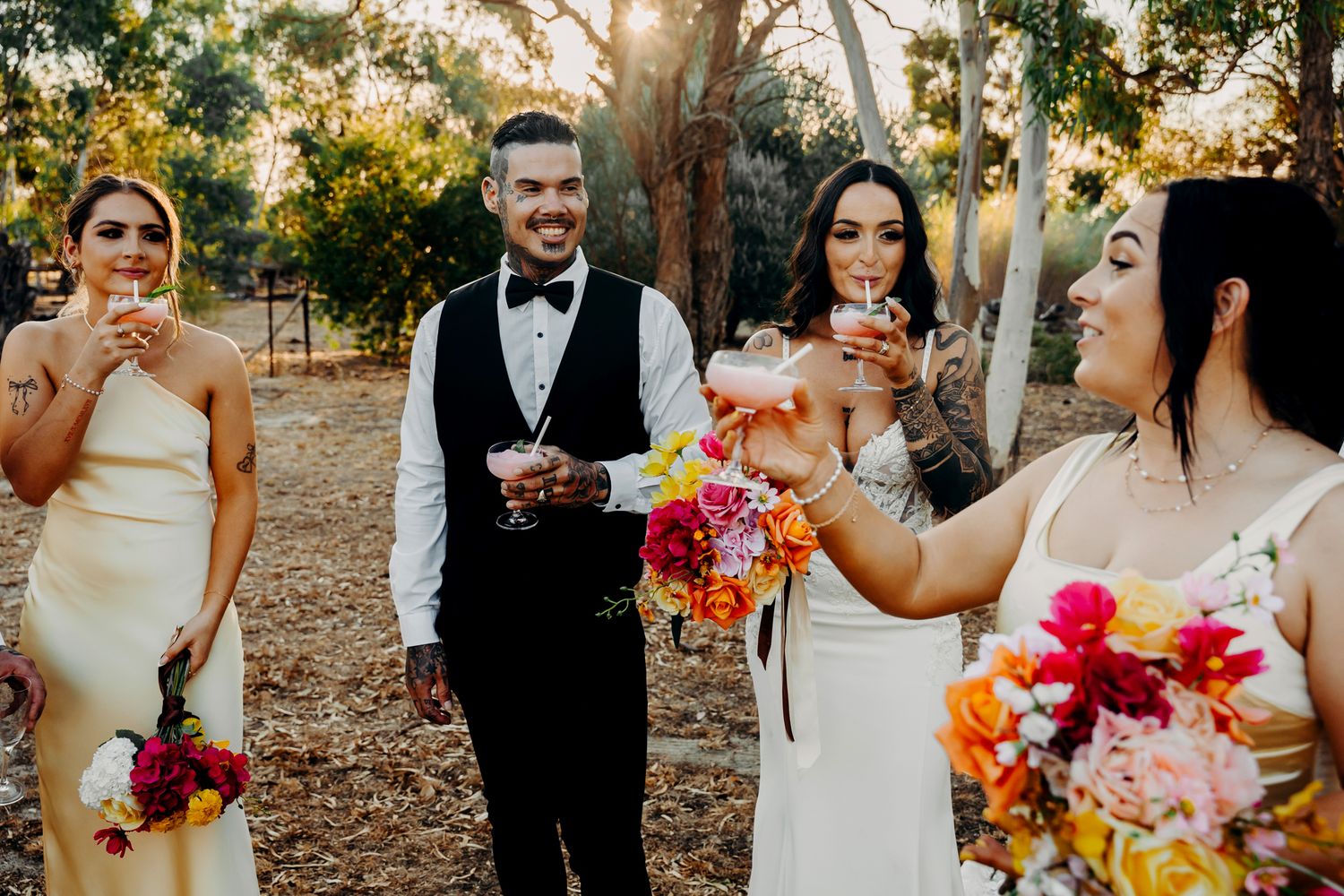 Wedding party celebrating outdoors with vibrant coral and pink floral bouquets in a rustic woodland setting.