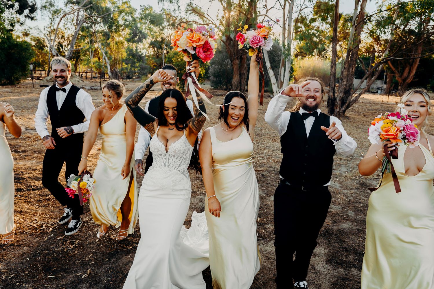 A joyful wedding party celebrating with raised bouquets in an outdoor wooded setting at sunset.
