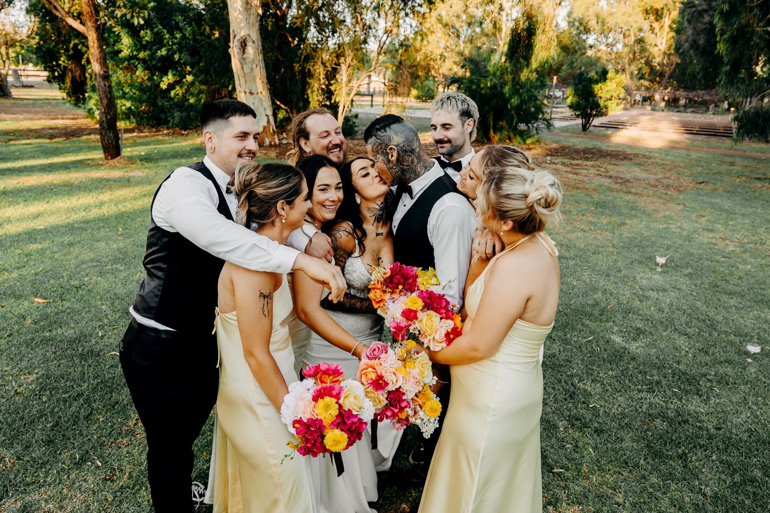 Wedding party gathers for a group photo holding vibrant pink and orange bouquets on a sunny lawn.