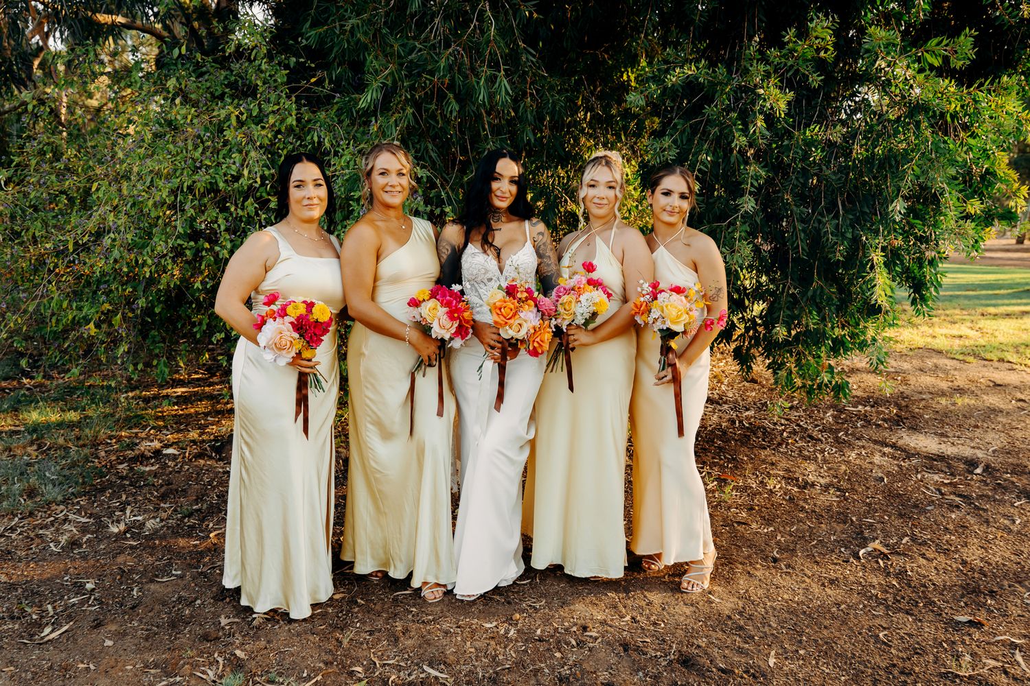 Bridesmaids in ivory floor-length dresses hold vibrant coral and pink bouquets while standing on a pathway outdoors.