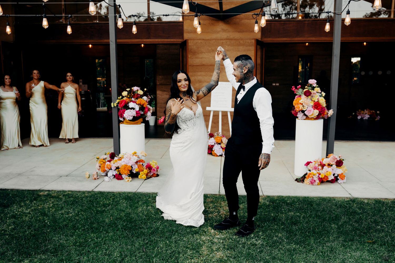 Wedding party gathered at an outdoor venue with string lights and decorative floral arrangements.
