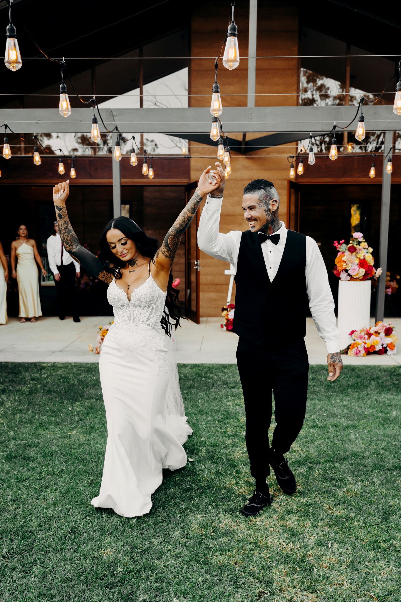 Newlyweds celebrate with raised arms under string lights at an outdoor reception venue.