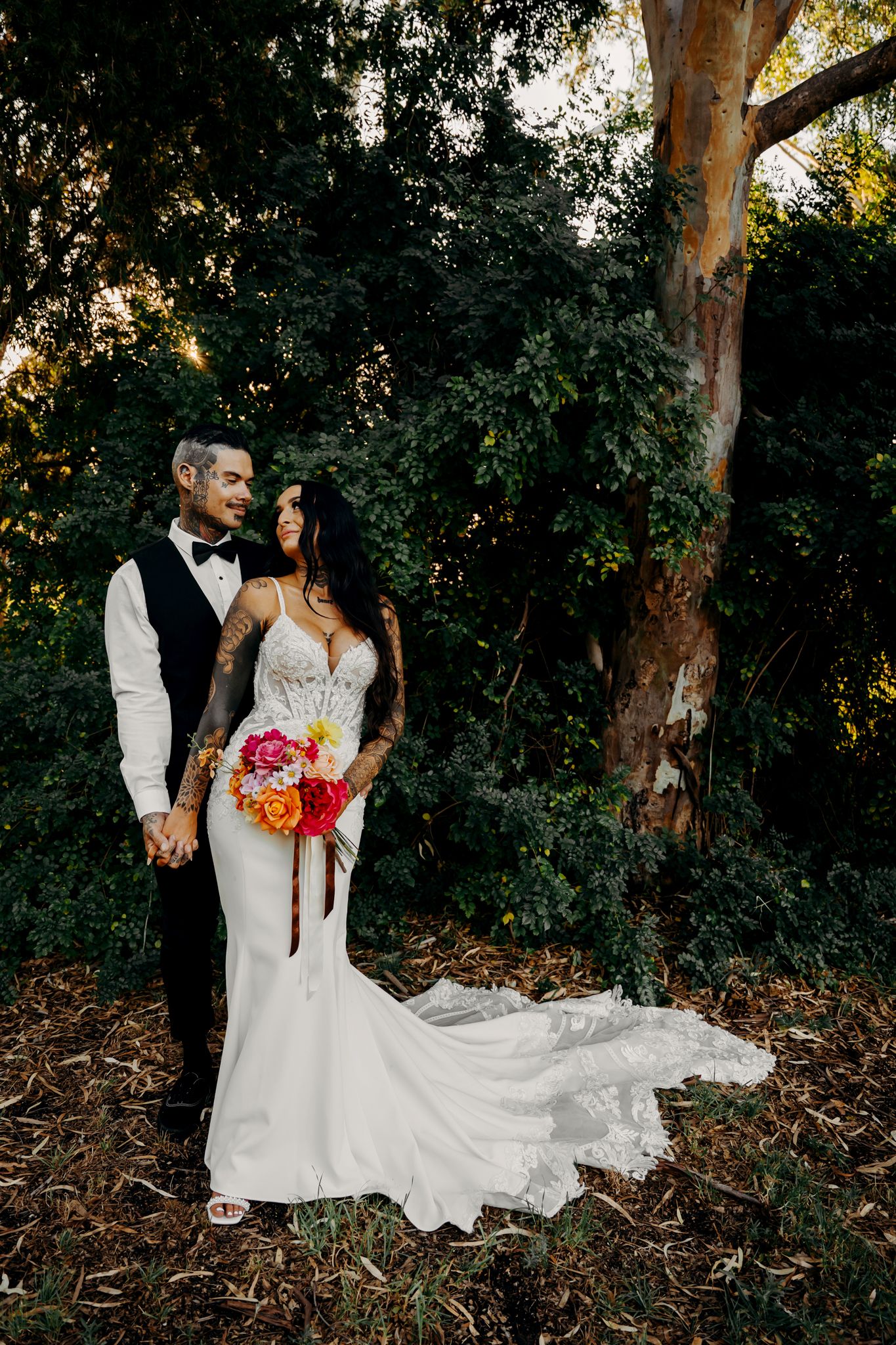 Couple in wedding attire hold hands beneath towering eucalyptus trees at sunset, featuring vibrant bouquet and flowing gown.