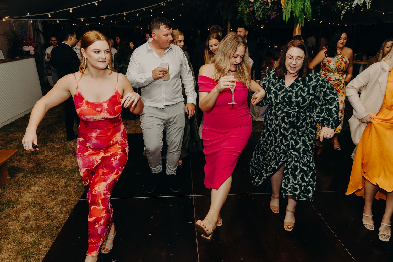 Wedding guests dancing in colorful dresses on dance floor at evening reception.