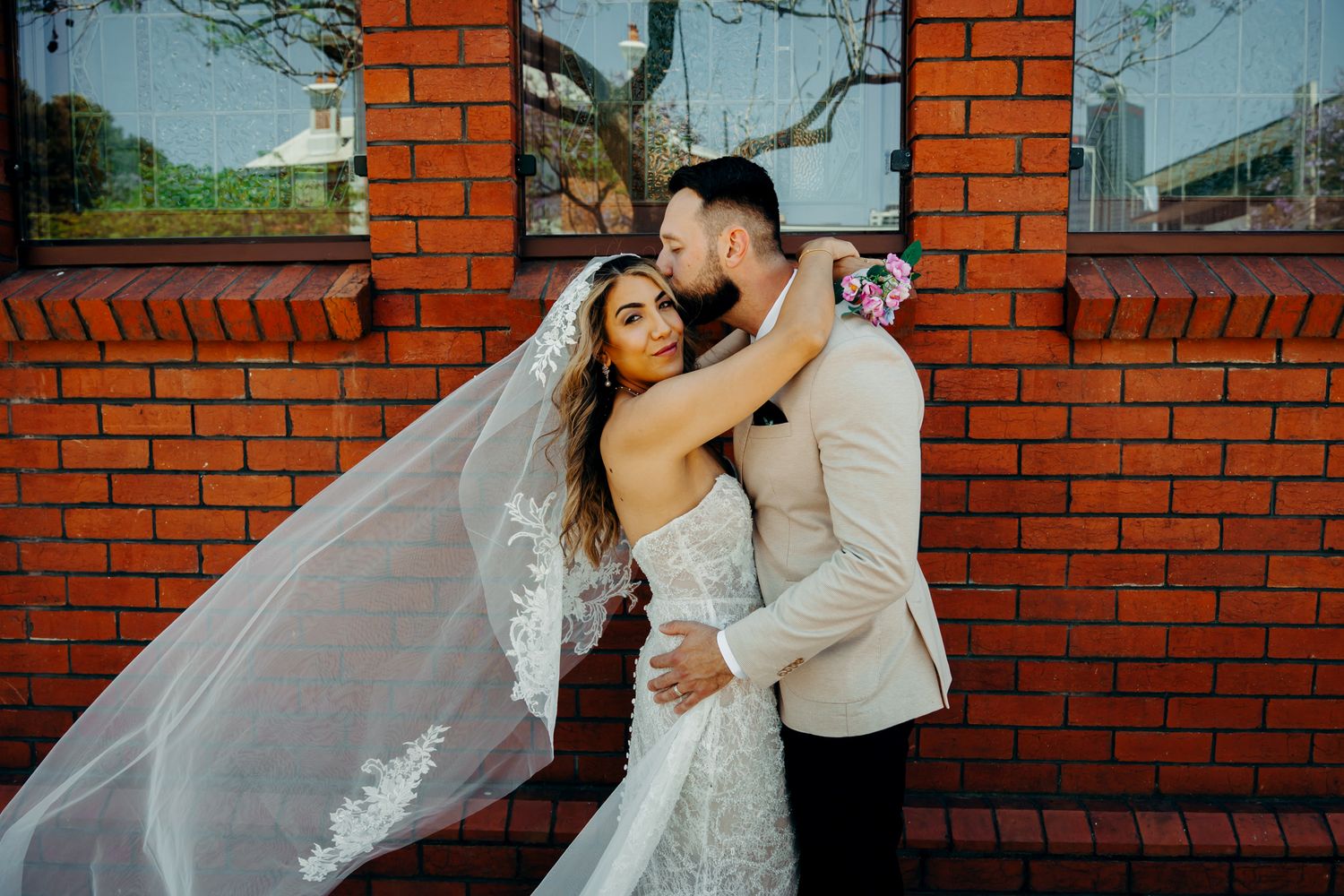 A romantic wedding couple shares tender moments against a red brick wall backdrop during their outdoor photoshoot.