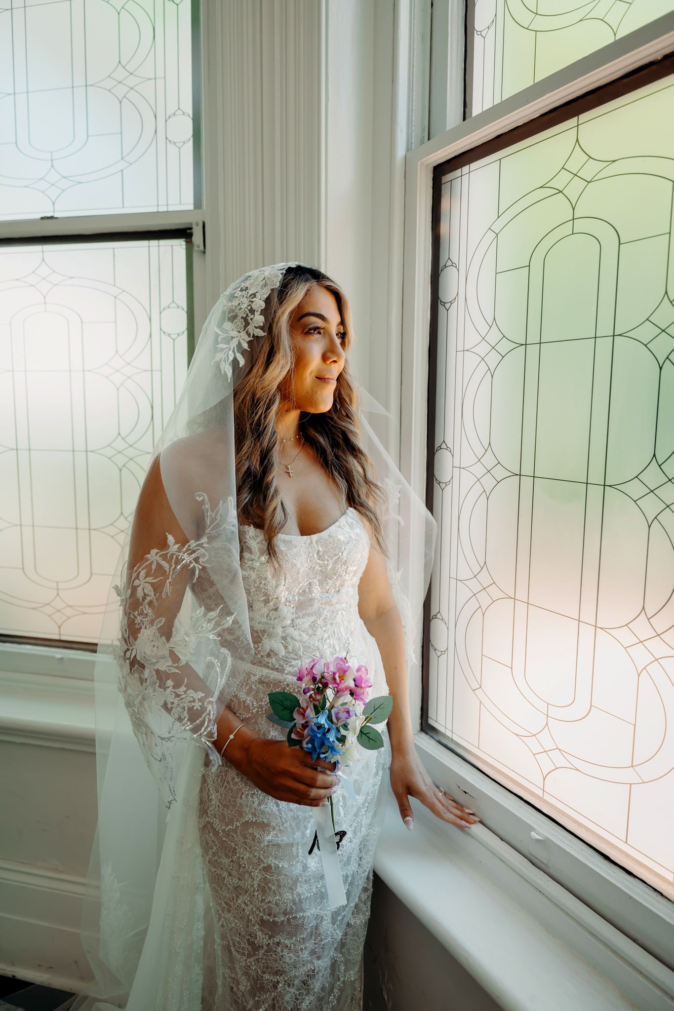 A bride in a white lace wedding dress and veil gazes out a window in a series of romantic getting-ready photos.