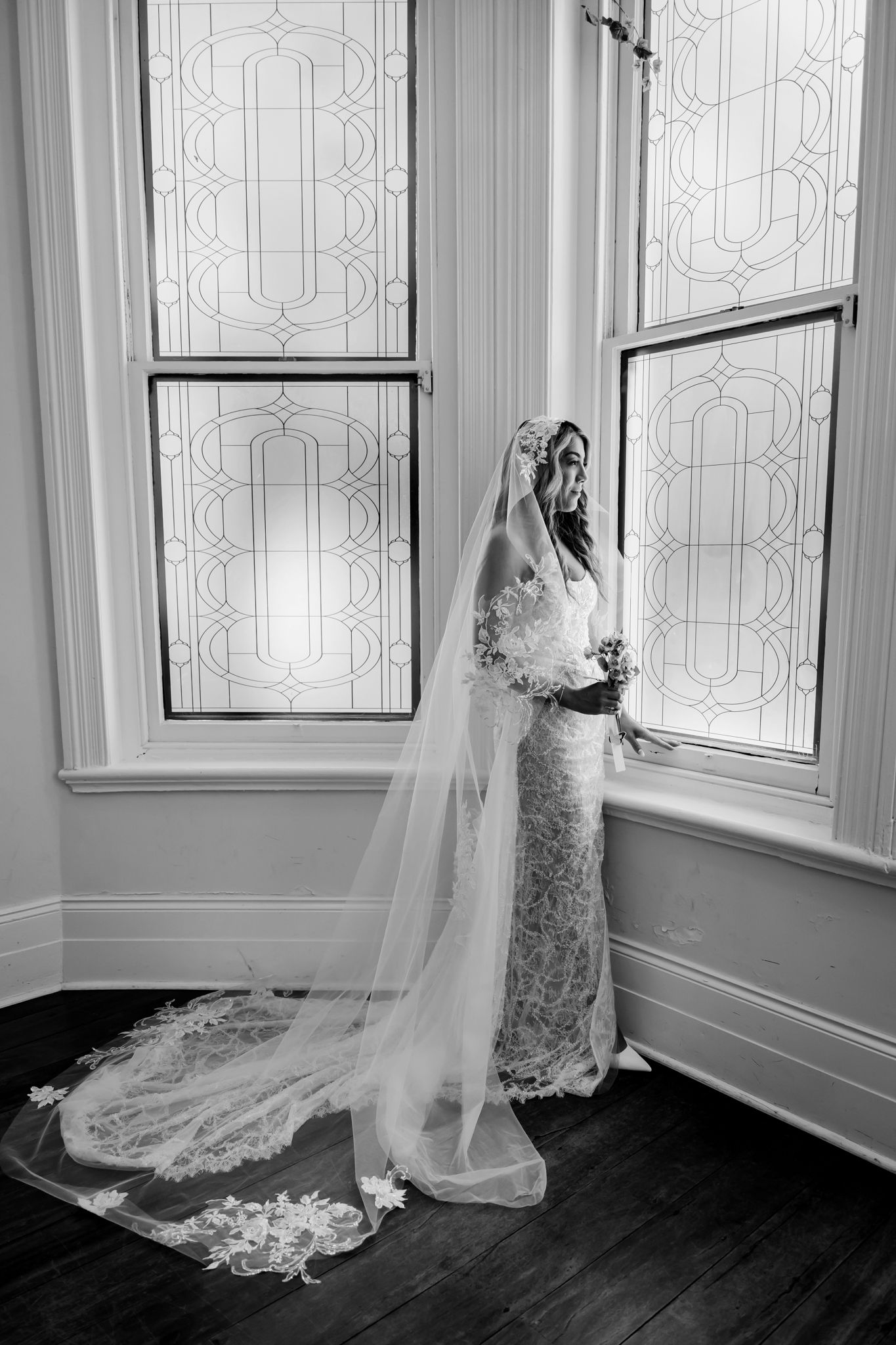 A bride in an elegant lace gown and flowing cathedral veil stands by ornate windows in a vintage room.