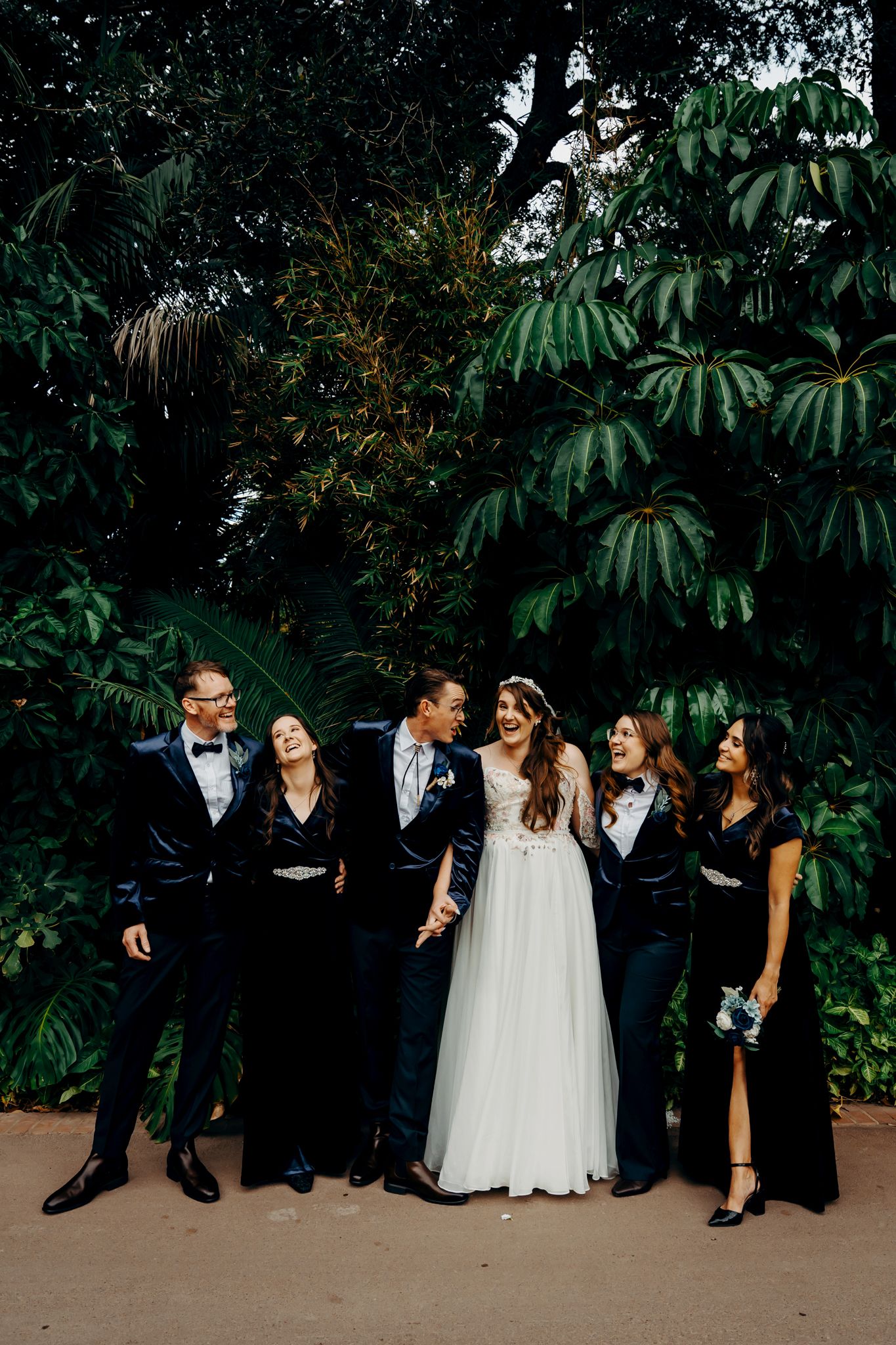 Wedding party poses together in elegant black attire and white gown against lush tropical garden backdrop.